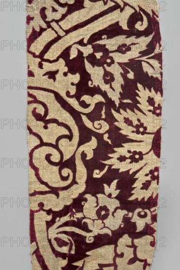 Fabric fragment, silk fabrics, fragments of patterned silk, chiseled velvet, Turkish fabric. Beige and gold pattern surrounded by cherry-cut fleece on a velvet background. Pattern features saz leaves with a fine floral ornament, lotus-shaped palmettes, and large, winding, forked, thorny flower stems. 17th century, Ottoman Empire 1299–1922, height 86 cm, width 25.9 cm., A narrow vertical textile fragment with a repeating ornate pattern in deep burgundy and a pale beige background, showing curved scrolls, stylized leaves, and floral motifs; the burgundy shapes form bold, velvety areas contrasted against the flatter beige ground, with irregular edges and small frayed threads along the sides, and the fabric surface exhibiting a slightly textured, plush appearance.