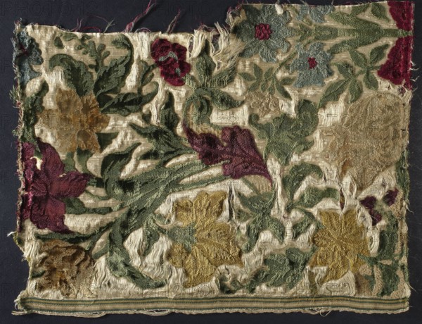 Fragment of patterned velvet fabric, 17th/18th century, Italy . Multi-colored chiseled cut and looped pile velvet on a cream satin weave ground cream silk warp and weft. Patterned velvet in which parts of the pattern have two pile heights—cut higher and looped lower—creating a contour effect. Colors: cherry red, burgundy, dusty yellow, honey-yellow, gray-blue, dark green. Design: symmetrical ; large, multi-flowered, densely leafed bouquets exceeding the width of the piece, depicting tulips, lilies, daffodils, roses; flower calyxes and leaves rendered in natural colors fiori naturali. Preserved silk weaving edge, width 1.6 cm, with green-beige-white-yellow stripes. Dimensions: height 40 cm, width 31.5 cm. Fragment of silk fabric;, A rectangular piece of frayed fabric with a floral pattern showing a group of leaves and flowers in shades of green, gold, burgundy, and muted blue on a worn cream background; the textile has areas of exposed threads and torn edges with a strip of woven edge i...