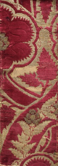Fabric fragment, chiseled velvet with plant ornament, patterned fabrics. Rectangular fragment of dark red velvet decorated with large plant motifs: winding stems and frayed leaves, a large five-petaled, and a smaller thistle-like flower. Pattern made of sheared and looped velvet and metal thread loops; metal thread in the background. 15th century, Italy. Height 97 cm, width 20 cm., A patterned textile with a rich red background and metallic gold woven areas forming stylized leaf and floral motifs; the red sections have a plush, velvety texture while the gold areas appear flatter and slightly shiny; the design shows curving outlines, serrated leaf edges, and a few raised embroidered clusters in a darker gold tone; there are some worn spots and loose threads where the fabric shows fraying and slight discoloration.