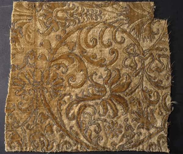 Fragment of ochre-colored sheared velvet with a raised arabesque pattern bordered with embroidery. Embroidery with gold-braided metal thread. Pattern design based on popular motifs from model books; a stylized spirally twisted tendril in the form of the head of a deeply bloomed flower matches a popular letter pattern book by Johann Theodor de Bry. 16th century, Italy. Fabrics, chiseled velvet, plant motifs. Height 28 cm, width 30 cm. Creator, provenance unknown., A square fabric fragment with frayed edges displayed against a dark background, showing a raised floral and leafy pattern in a velvety texture; the fabric is primarily golden-beige with darker golden-brown areas forming stylized flowers, petals, and curling vines, and there are thin metallic threads outlining many of the motifs that appear silver-gray; the surface shows variations in sheen and pile, with crushed and smooth patches creating light and dark contrasts across the decorative design.