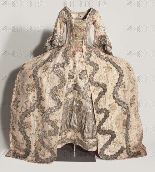 Robe à la française robe à paniers à coudes, two-piece, ca. 1764. Pink grosgrain with swirling silver-lamellar brocaded pattern, silver lace ribbons and bouquets of flowers; lace ornaments and ribbon. Outer dress manteau open at the front with a pleated train at the back. Bodice stiffened with canvas and boning 22, laced at front and back under the pleats. Elbow-length sleeves cut in a curved pattern with double flounces trimmed in silver lace. Dress made of eight panels; quadruple pleats at the back preserve full fabric width; twenty-two long bonings extend beyond the bodice line. Silver lace appliqué on edges echoes the wavy brocaded pattern. Apron of same fabric, hemmed with a wide ruffle and silver lace appliqués repeating the wavy motif. Worn on an oval cornea. Fabric report: sample height 37 cm x width 27 cm; fabric width 55.5 cm. Garment length 167 cm; width at bottom of manteau 430 cm., Poland use; France fabric., A historical-style dress displayed on a stand with a fitted bodi...