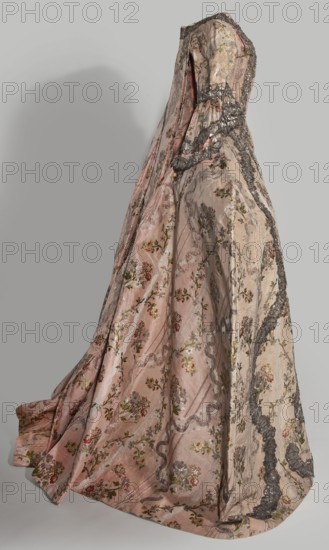 Robe à la française robe à paniers à coudes, two-piece, 1764. Pink grosgrain with swirling silver-lamellar brocade, silver lace ribbons and bouquets of flowers; lace ornaments and ribbon. Outer dress manteau open at the front with a pleated train at the back; bodice stiffened with 22 bonings, laced at front and back under the pleats. Elbow-length curved sleeves finished with double flounces trimmed with silver lace. Dress cut in eight panels; quadruple pleats at the back maintain full fabric width, with cuts and fittings at the front of the bodice. Twenty-two long bonings extend beyond the bodice line. Silver lace appliqué on the edges repeats the wavy rhythm of the brocaded pattern. Apron of same fabric, hemmed with a wide ruffle and matching silver lace appliqués. Worn on an oval cornea. Fabric: height 37 cm x width 27 cm; fabric width 55.5 cm. Garment length 167 cm; width at bottom of manteau 430 cm., Poland use; France fabric. Women’s fashion, 18th century., A long, ornate gown dis...