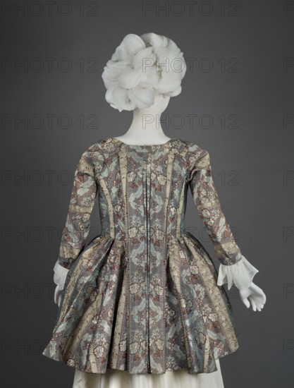 Top of the cornea, floral patterns, clothing, women's clothing, patterned fabrics, fashion, 18th century clothing, 1770s, Silesia, France fabric, clothing, length 85 cm, width 298 cm bottom, A back view of a figure wearing a patterned dress with a fitted bodice and a pleated skirt, seen against a plain dark gray background. The dress fabric shows an ornate floral and scroll design in muted tones of blue, pale green, beige, brown, and hints of ivory, with vertical panels and pleats at the waist creating a full skirt silhouette. The long sleeves end in sheer white ruffled cuffs. The visible head is covered by a sculpted, voluminous white headdress with layered, rounded folds. The hands are white and gloved or sculpted, positioned at the sides.