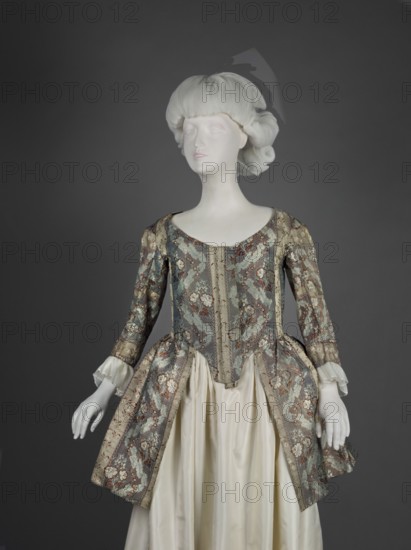 Top of the cornea, floral patterns, Henryków German: Heinrichau, clothing, women's clothing, patterned fabrics, fashion, 18th century clothing, 1770s, Silesia, France fabric, clothing, length 85 cm, width 298 cm bottom, A sculpted mannequin with a pale white head and hands wears an ornate historical dress; the bodice and overskirt are patterned in muted teal, gold, brown, and cream with floral motifs and metallic sheen, and the underskirt is a smooth ivory satin. The mannequin's hair is a sculpted pale white with soft waves, and the background is a solid dark gray. The sleeve cuffs show a ruffled cream fabric, and the overall silhouette includes a fitted waist with a flared, pleated overskirt.