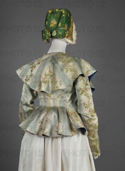 Women's caraco women's bodice, 1770s, Europe. Cream and blue silk brocaded with rose branches and colorful floral patterns; patterned fabric embellished with brocade. Straight-cut front fastened with hooks, slightly overlapping lapels; low neckline finished with a large turn-down semicircular collar covering the shoulders. Long set-in narrow sleeves, cut in a curve at elbow height. Back fitted at the waist, widened below by triangular gussets creating a generous basque/ruffle. Dark blue canvas lining; inside under the flaps a wide red canvas belt is sewn on and laced at the front; lace-up waistband at the lining. Length 56.5 cm; bottom circumference 249 cm. Maker, attributed to Brandt, Józef 1841–1915 noted in provenance., A mannequin shown from behind wears a fitted jacket with a peplum and a wide, cape-like collar, the fabric displaying pale blue and light beige vertical stripes with floral motifs in green, yellow, and pink; the jacket sleeves carry similar floral embroidery and a da...