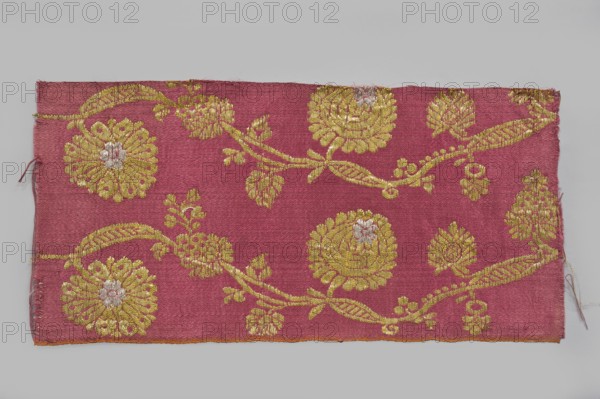 Fabric fragment, fragments of silk patterned semi-silk fabric with a red background in a satin weave and brocaded decoration using two types of braided metal thread with yellow and white cores. Vertically arranged pattern of a waving, fanciful flowered and leafy branch with small leaves and large oval heads of stylized pompon flowers alternating in horizontal rows. Dyes: indigo, brazilwood. Semi-silk satin, brocaded. Weaving edge: 0.3 cm wide, orange cotton warp and cherry cotton weft in plain weave. Construction: satin warp weave; thicker red cotton wefts and red silk warp giving stiffness. Pattern introduced with brocade wefts using two types of loosely braided threads – a gold plate on a yellow cotton core and a silver plate on a white cotton core. Continuous longitudinal pattern on a red background of symmetrical trails of bending gold twigs with leaves, fruit, and two types of flower heads with silver centers. Pattern repeat width 10.5 cm. Selvedge width 0.3–0.4 cm. I-thread: yell...