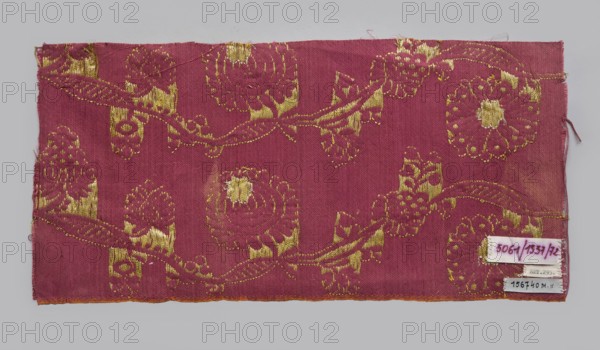 Fabric fragment, fragments of silk patterned fabric with plant motifs, indigo and Brazilwood dyes. Semi-silk with a red satin-weave background and brocaded decoration using two types of braided metal thread with yellow and white cores. The vertically arranged pattern shows waving, fanciful flowered and leafy branches covered with small leaves and large oval stylized flower heads alternating left and right in horizontal rows referred to as pomponblüten. Floated and brocaded with double metal thread gold plate on yellow core and silver plate on white core. Continuous pattern along the length on a red background—symmetrical trails of regularly bending gold twigs with leaves, fruit, and two types of flower heads with silver centers. Construction: satin-warp weave; thicker red cotton wefts and red silk warps give stiffness. Weaving edge 0.3–0.4 cm wide with an orange cotton warp and cherry cotton weft in plain weave. Pattern repeat c. 10.5 cm. Tapestry-like brocaded satin, likely 17th centu...