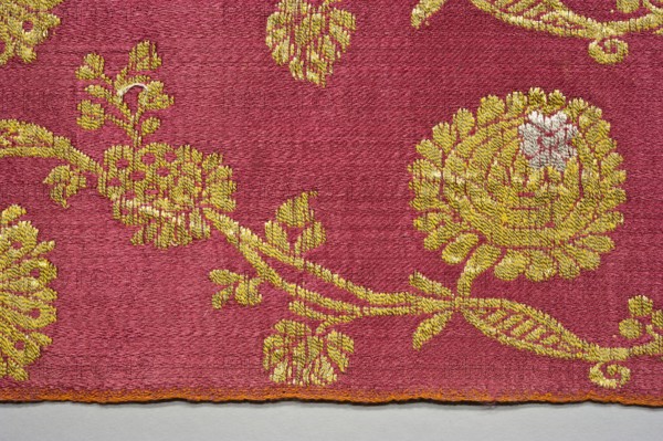 Fabric fragment, fragments of silk patterned fabric with plant motifs, indigo and brazilwood dyes. Semi-silk with a red satin-weave background and brocaded decoration using two types of braided metal thread with yellow and white cores. Vertically arranged pattern of waving, fanciful flowered and leafy branches with small leaves and large oval stylized flower heads alternating horizontally; called pomponblüten pompon flowers. Similar fabrics dated to the second quarter of the 17th century. Floated and brocaded with double metal thread with a yellow silk core; pattern report width c. 10.5 cm. Weaving edge c. 0.3–0.4 cm wide, orange cotton warp and cherry cotton weft in plain weave. Construction: satin warp weave; thicker red cotton wefts and red silk warp give stiffness. Brocade wefts use two types of loosely braided threads: gold plate on a yellow cotton core and silver plate on a white cotton core. Continuous longitudinal pattern on a red background—symmetrical trails of bending gold t...