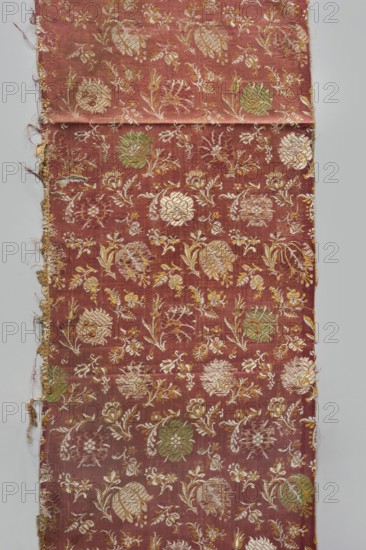 Fabric fragment, fragments of silk patterned fabric, 17th century, Italy . Warp-woven satin background with pale red silk warp and two alternating wefts white and yellow creating the pattern using the lisère technique—floating runs in diagonal, fairly wide bands. Brooching wefts: green and beige. High fabric tightness. SATIN WITH A TWO-COLOR WEFT, LISÉRE. Pattern repeat 29 x 17.5 cm. Floral motifs in three types arranged at close intervals: two kinds of carnations rows facing left or right, tulips with drooping calyxes, and roses with spread petals, with smaller flower projections scattered between them, facing in different directions. Colors of flowers and elements variable—white and yellow stems and leaves, green and beige calyxes. Height 112 cm, width 28.5 cm., A long rectangular piece of fabric with a muted reddish-brown background, patterned with a repeating floral design in pale beige, gold, and green tones, showing stylized blossoms, leaves, and curling stems; the fabric has fra...