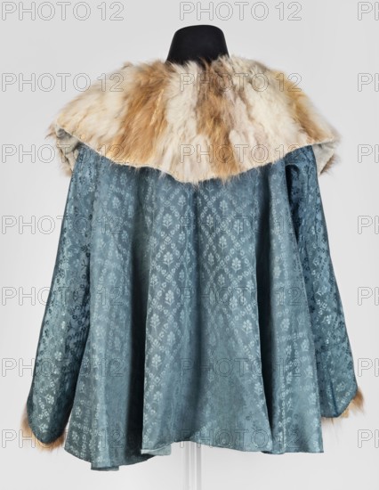 Jubka, long women's caftan jubka, hip-length, narrow in the shoulders, flared at the bottom; late 18th century, Poland. Large turn-down collar entirely trimmed with fur two types of fur decoratively connected in narrow bands. Long set-in sleeves, narrow at the top, flared at the bottom, lined with white sheepskin with strongly twisted fleece. Geometric ornaments; women's clothing, fashion. Dimensions: length 63 cm, circumference 292 cm at the bottom., A garment displayed on a black mannequin with a wide fur collar in shades of cream, beige, and light brown, draped over a flowing fabric body in a muted teal-blue with a subtle floral damask pattern, the sleeves and hem showing the same teal-blue fabric and small touches of the light brown fur at the cuffs, all against a light gray background.