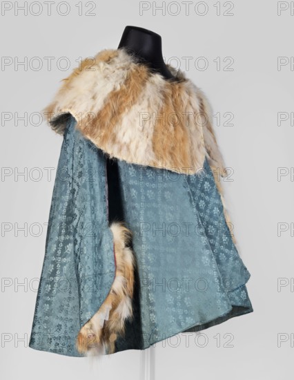 Jubka, long women's caftan jubka, hip-length, narrow in the shoulders, flared at the bottom. Large turn-down collar, entirely trimmed with fur two types of fur decoratively connected in narrow bands. Long set-in sleeves, narrow at the top, flared at the bottom, lined with white sheepskin with strongly twisted fleece. Geometric ornaments. Late 18th century, Poland. Clothing; length 63 cm, circumference 292 cm at the bottom., A mannequin displays a cape with a large fur collar and fur trim; the fur is a mix of cream, beige, and light brown tones, appearing fluffy and soft, while the main fabric of the cape is a blue-green color with a subtle floral or damask woven pattern visible across its surface; the interior edge and some trim also show the same cream and light brown fur, and the mannequin head is black against a pale gray background.