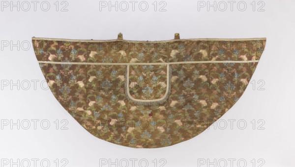 Church cope made of silk brocaded with colorful threads green, cream, blue-pink, cherry, 18th century, France; decorated with flowers, leaves, and plant ornaments; liturgical vestment church paraments, used in sacred art for churches and chapels; height 137 cm, width 302.5 cm., A semicircular textile with a patterned surface in brown, green, blue, pink, cream, and gold tones, featuring a central U-shaped flap edged with a beige fringe and trim, a horizontal beige band near the top, two small fabric loops along the upper edge, and a repeating floral or botanical motif across the entire piece against a pale neutral background.