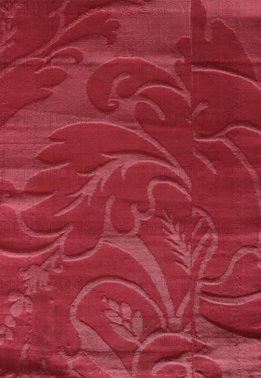 Fabric fragment, silk damask, patterned silk with floral motifs. Fragment of cherry damask with a large, likely symmetrical pattern featuring stylized acanthus leaves. Silk intended for upholstery. 19th century, France, height 66 cm, width 22.5 cm., The image shows a textured fabric with a raised floral and leaf pattern in shades of red, featuring darker and lighter red areas that create a subtle sheen; swirling petal and leaf shapes form a repeating ornate design across the surface, with visible fabric weave and slight horizontal creases and seams.