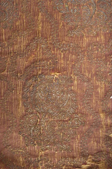 Fabric fragment, silk brocade grosgrain, 1st half of the 17th century, Italy. On a red background a gold floral pattern of projections on a curved leafy stem with a large stylized tulip calyx; motifs arranged in rows shifted by half a repeat, alternating lean left and right, with a wavy twig with small leaves between them. Construction: yellow silk weft, red silk warp in grosgrain weave; brocaded thread of gold plate braiding on a yellow silk base. Pattern repeat: 33 cm x 17 cm. Border width: 1 cm. Colors: yellow-brown and gold threads. Dimensions: 39.5 cm high x 43 cm wide., The image shows a textured fabric with an intricate woven pattern featuring floral and swirling motifs. The dominant colors visible are reddish-brown, gold, and muted green, with highlights of pale gold or beige creating a slightly metallic sheen. The surface displays a mix of vertical striations and raised embroidered-like areas where the pattern is more pronounced, and there are a few small worn spots where the ...