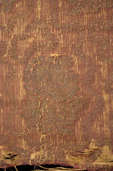 Fabric fragment, silk brocade brocaded grosgrain, 1st half of the 17th century, Italy. Red ground with a gold floral pattern of projections on curved leafy stems, featuring a large stylized tulip calyx. Motifs arranged in offset rows shifted by half a repeat, alternating lean left and right; between them a wavy twig with small leaves. Construction: yellow silk weft, red silk warp in a grosgrain weave; brocaded thread of gold plate braid on a yellow silk ground. Pattern repeat: 33 cm × 17 cm. Border width: 1 cm. Colors: yellow-brown, gold. Overall dimensions: height 39.5 cm, width 43 cm., A close-up of a woven textile showing a worn patterned surface with a vertical striated background and an overlaid ornate floral motif; the fabric displays frayed edges and threadbare areas near the bottom, with visible loose threads and small gaps. The colors visible are shades of reddish-brown, gold, and muted olive, with lighter golden highlights in the raised embroidery and darker brownish-red thre...