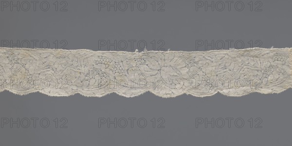 Lace fragment, early 18th century, Binche. Materials: lace fabric; dimensions: width 3 cm, length 130.5 cm. Design: plant ornaments with twig motif featuring large leaves and flowers roses, rosettes; upper straight edge with half stars containing rosettes; lower edge gently serrated; varied background. Creator: unknown., A horizontal strip of lace with an intricate floral and scrollwork pattern stretches across a gray background; the lace appears off-white to cream with subtle darker beige discolorations in places, featuring a scalloped lower edge, delicate openwork, and a few frayed threads along its top edge.