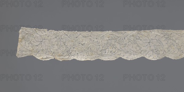 Lace fragment, early 18th century, Binche. Plant ornaments with rosette and rose motifs; twig motif with large leaves and flowers. Upper straight edge has half stars with rosettes inside; lower edge is gently serrated. Background varied. Lace fabric; width 3 cm, length 130.5 cm., A horizontal strip of delicate lace with a scalloped lower edge stretches across a gray background; the lace is off-white with small areas of light tan discoloration, featuring an intricate openwork pattern of swirling floral and leaf-like motifs, fine looping threads, and tiny holes, with slightly irregular edges and a few loose fibers visible.