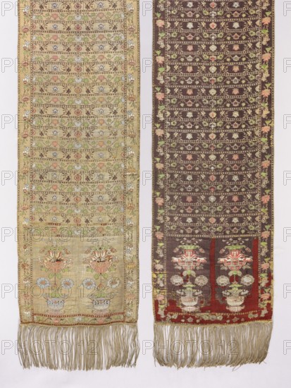 Kontusz belt, Daniel Chmielewski's Studio Kraków; belt manufacturer; 1796–1825. Solid, double-sided kontusz belt woven ca. 1794–1796, Kraków. Length 372 cm, width 37 cm. Weave density approx. 19/32 x2. Golden fringe sewn on. Ornament: - Heads: two motifs of flower bouquets placed in wide vase forms resembling amphoras. - Center: transverse shelf with two types of floral tendrils. - Edges: vertical and horizontal tendrils like on shelves. - Borders: rhombuses and ovals. Colors: - Right side: gold background with brown, green, blue, red and white ornament. - Left side: brown background with gold ornament and the same colors as the right., Two long woven textiles hang side by side against a pale background, each with decorative fringed bottoms. The textile on the left is a light beige color with horizontal bands of floral motifs in soft pastels including pale blue, pink, green, and muted brown, and a narrow floral border along its edges; the lower section shows larger stylized floral arra...