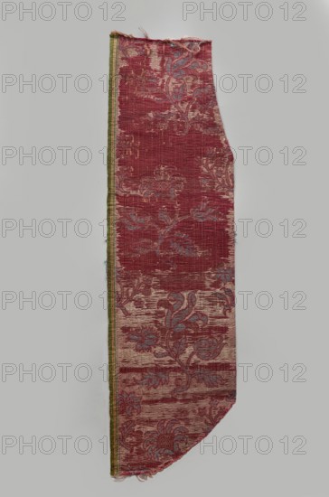 Fragment of fabric, silks, brocade, floral and plant patterns, double-warp brocade. On a white and silver background, blue motifs with a cherry outline. On a white and cherry background, densely placed motifs of blue flower bushes with a cherry outline — sunflower-like flowers on leafy bent stems, alternating in rows shifted by half the motif width, and S-shaped flower branches with a tulip and a carnation. Fragmentary elements suggest additional smaller motifs between the two main ones. Construction: background in plain warp weave - basic linen thread, bold cherry-red silk warp; additional pink silk warp binds in a twill weave the patterning threads: silk blue pattern and white background covering, plus bundles of two silver lamellas running on the surface across the fabric width. Pattern repeat: length 45 cm. Weaving edge: 1.3 cm wide in green with yellow, pink and white stripes. Date: 16th/17th century, Italy. Dimensions: height 77 cm, width 21.5 cm., A long, narrow textile scrap wi...