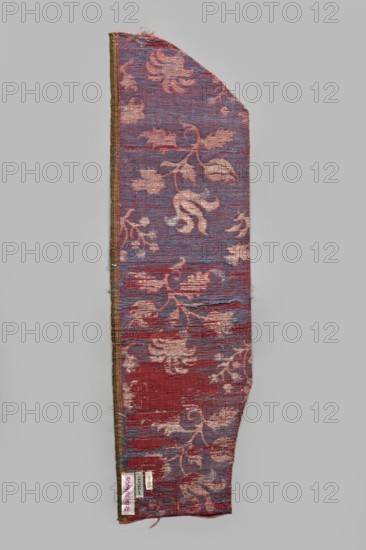 Fragment of fabric, silks, patterned brocade with floral and plant motifs, double-warp brocade. On a white and silver background, blue motifs with a cherry outline. On a white and cherry background, densely placed motifs of blue flower bushes with a cherry outline—sunflower-like flowers on leafy, bent stems, alternating in rows shifted by half the motif width, and S-shaped flower branches with a tulip and a carnation. Fragmentary elements suggest additional smaller motifs between the two main ones. Construction: background in a plain warp weave of basic linen thread; bold cherry-red silk warp; additional pink silk warp binding in a twill weave the pattern threads: silk blue pattern and white background covering; bundles of two silver lamellas running on the surface across the fabric width. Pattern repeat length 45 cm. Weaving edge 1.3 cm wide in green with yellow, pink and white stripes. 16th/17th century, Italy; textiles; height 77 cm, width 21.5 cm., A long narrow textile fragment wi...