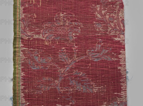 Fragment of fabric, silks, patterned brocade with floral and plant motifs, double-warp brocade, 16th/17th century, Italy. On a white and silver background, blue motifs with a cherry outline. On a white and cherry background, densely placed motifs of blue flower bushes with a cherry outline: sunflower-like flowers on a leafy bent stem, alternating in rows shifted by half the motif width, and S-shaped flower branches with a tulip and a carnation; fragmentary elements suggest additional smaller motifs between the two main ones. Construction: background in a plain warp weave of basic linen thread with bold cherry-red silk warp; additional pink silk warp binds in a twill weave the pattern threads: silk blue pattern and white background covering, plus bundles of two silver lamellas running on the surface across the full width. Pattern repeat length: 45 cm. Weaving edge: 1.3 cm wide in green with yellow, pink and white stripes. Dimensions: height 77 cm, width 21.5 cm., A worn woven textile fr...