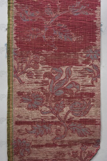 Fragment of fabric, fabrics, silks, fabric with preserved selvedge, patterned brocade, floral and plant patterns, double-warp brocade. On a white and silver background, blue motifs with a cherry outline. On a white and cherry background, densely placed motifs of blue flower bushes with a cherry outline — sunflower-like flowers on a leafy bent stem, alternating in rows shifted by half the motif width, and S-shaped flower branches with a tulip and a carnation. Fragmentary elements suggest additional smaller motifs between the two main ones. Construction: background in a plain warp weave — basic linen thread, bold cherry-red silk warp; additional pink silk warp binds in a twill weave the pattern threads: silk blue pattern and white background covering, plus bundles of two silver lamellas running on the surface across the fabric width. Pattern repeat length 45 cm. Weaving edge 1.3 cm wide in green with yellow, pink and white stripes. Renaissance/Baroque, 16th–17th century, Italy. Textiles;...