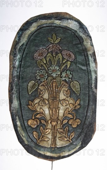 Fragment of a cordovan armrest, cordovan leather, flowers, flower bouquets, furniture upholstery. Oval-shaped leather armrest with embossed, painted, silver-plated, and gilded decoration of a bouquet of flowers in a vase; the vase is symmetrically decorated on both sides with S-shaped leafy branches. Colors: turquoise-green background, gold branches, pale pink, cream, and turquoise flowers with light green leaves. 18th century, Europe. Height 36 cm, width 21 cm., An oval-shaped carved panel with a raised floral design showing a stylized bouquet in a vase; the panel surface is predominantly dark green with areas of brown and muted olive, the carved floral elements and vase are in shades of tan, gold, and brown, and some flower petals and leaves show pale green and muted bluish-green highlights; the oval border has layered tones of dark green, bluish-gray, and brown with lighter worn streaks, and the background around the bouquet is a deep green with visible textured cracking and aged su...