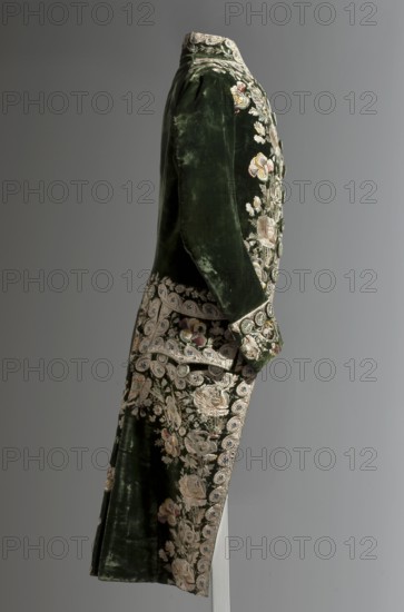 Green velvet tailcoat, embroidered with motifs of field and garden flowers — roses, cornflowers, chamomiles, and bluebells. Fitted at the top, widened at the back from the waist by slit pleats. The flaps at the top, cut in a semicircle, overlap at the bust, and are cut short at the bottom. Fastened with hooks at breast level. Long sleeves, cut in a curve at elbow height, set in, and finished with cuffs. High, stand-up collar. Back cut with a center seam. The pleats are slit at the back, not overlapping in the center. Rich embroidery covers the edges of the pockets, pocket flaps, cuffs, and collar. Fabric-covered and embroidered buttons. Green sheared silk. Embroidery with twisted and untwisted silk yarn light pastel shades with a predominance of white, satin lay stitch, white satin appliqué. Lining of cream plain silk. Hand-sewn, early 19th century, Europe. Garment; length 111 cm., A side view of a dark green velvet coat with ornate floral embroidery, showing a high collar, long fitted...