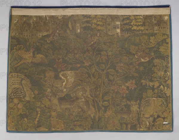 Tapestry-verdure, verdure with flowers and leaves, floral ornaments, 17th century, Flanders; fabric; height 194 cm, width 240 cm., The image shows a woven tapestry with a predominantly muted green background and a border in blue and beige; the tapestry features a complex botanical scene with a group of leafy vines and several stylized flowers in shades of pink, red, and yellow; animal figures appear throughout, including birdlike shapes and other creatures rendered in brown, tan, and gold tones with patterned feathers and fur; the upper edge displays architectural silhouettes in faded beige and brown, and there are areas of wear and lighter thread revealing underlying canvas in pale beige; overall the palette includes green, brown, tan, gold, beige, blue, pink, and red.