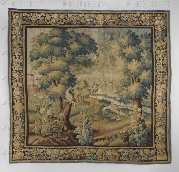 Tapestry-verdure, In the foreground, three flowering shrubs; behind them two birds, one perched on a tree trunk, and a bird fluttering between the branches. Two large trees frame the scene. In the background, a landscape with architecture and a river. Border with motifs of flower bouquets in vases. Flowers, leaves, floral ornaments, trees, birds, landscapes, borders. Colors: green, sand, sky blue, and dark blue. 17th century, Flanders. Woven fabric; height 272 cm, width 278 cm., A woven tapestry showing a pastoral scene framed by an ornate decorative border; within the central field there are a few large leafy trees with foliage in shades of green, blue-green, and yellow, a winding stream or river in pale blue and off-white, grassy and earthy ground in shades of brown, tan, and green, and a group of plants with broad leaves in muted greens and bluish tones. Two birds appear in the foreground perched on or near tree trunks with plumage in pale cream, gray, and muted brown, and another b...