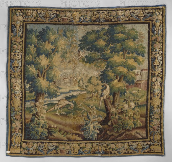 Tapestry-verdure, In the foreground, three flowering shrubs; behind them two birds, one perched on a tree trunk and one fluttering between the branches; two large trees. In the background, a landscape with architecture and a river. Border with motifs of flower bouquets in vases. Floral ornaments, flowers, leaves, trees, birds, landscapes, borders. Colors: green, sand, sky blue, dark blue. 17th century, Flanders. Woven fabric; height 272 cm, width 278 cm., A woven tapestry showing a landscape scene with a group of large leafy trees and a few smaller plants in the foreground, rendered in shades of green, gold, brown, blue, and cream. Near the center foreground, a bird perches on a tree stump while another bird feeds on the ground among grassy mounds and foliage in green and brown tones. In the midground a flowing waterway winds past grassy banks toward a distant area with indistinct built structures partially obscured by trees. The tapestry has a decorative border featuring floral and fo...