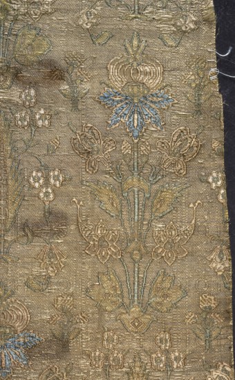 Fabric fragment, silk, fragments of patterned floral/plant motifs. Stylized plant motifs on a silver-green background in a symmetrical arrangement: flowering trees and lilies irises combining several flower species with small lanceolate leaves and scalloped edges. Brocade colors include blue, pale brown, pink, yellow, light green, and white. Printed area 26.5 x 15 cm; overall height 63.5 cm, width 17.6 cm. 17th century, Iran., A piece of woven fabric with an ornate floral pattern in shades of gold, beige, muted blue, and pale green, showing stylized flowers and leaves arranged vertically; the textile has a textured, slightly worn surface with visible threads and a darker edge along one side, and several areas where the weave appears frayed or disrupted.