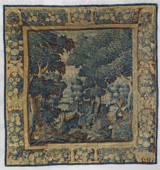 Tapestry-verdure, In the foreground are large trees and blooming shrubs and flowers; further on a river; beyond the river are rural buildings. Border: side panels with vases of flowers; horizontal panels with flower garlands; center with vases of flowers. Colors: sand, brown, blue, green, and ochre. 17th century, Netherlands. Woolen decorative fabric tapestry, height 319 cm, width 308 cm., A woven textile with an ornate central scene framed by an elaborate border, showing a landscape of densely packed trees and foliage in shades of blue, green, brown, beige, and muted gold; the central area features a group of large leafy trees, shrubs, and ground plants with detailed layered leaves and rounded canopies, along with a few rocky or root-like forms and low vegetation in darker blues and browns, while the surrounding wide border displays repeating floral and foliate motifs in beige, gold, blue, and brown set against a neutral background.