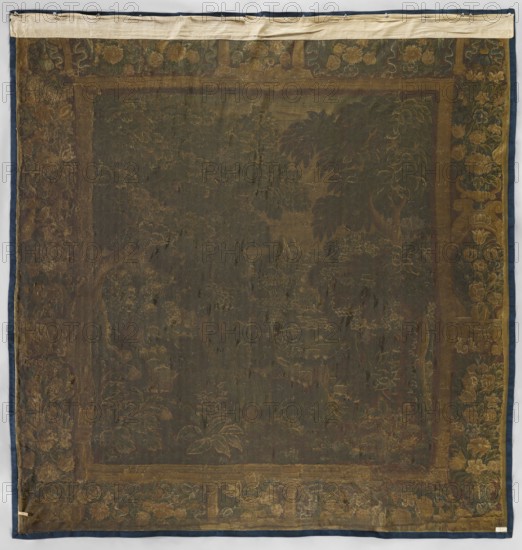 Tapestry-verdure, In the foreground are large trees and blooming shrubs and flowers; further on a river; beyond the river are rural buildings. Border: side panels with vases of flowers; horizontal panels with flower garlands; center with vases of flowers. Colors: sand, brown, blue, green, and ochre. 17th century, Netherlands. Woven decorative fabric tapestry, height 319 cm, width 308 cm., A large square textile with a dark central field surrounded by a patterned border; the fabric shows a faded floral and foliage design with leaves and flower shapes rendered in muted gold and brown tones against a deep greenish-brown background, and a surrounding band featuring floral motifs in shades of gold, olive, and dark green with touches of brown; the outermost edge is trimmed in a dark blue fabric and the top edge has a lighter beige strip attached; the surface appears worn and uneven in texture with areas of lighter and darker discoloration and visible weave.