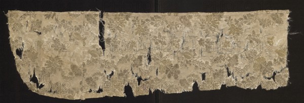 Fragment of silk fabric with a pattern of plant tendrils on a crimson background, pink and yellow floral/plant pattern; 17th century, France; height 80 cm, width 23.5 cm., A long, tattered strip of woven fabric displayed against a dark background, showing a floral damask pattern with raised motifs and many irregular tears and frayed edges; the fabric appears in shades of beige, cream, light gold, and pale olive with areas of darker brownish-gold in the patterned sections, and there are visible loose threads and holes throughout.