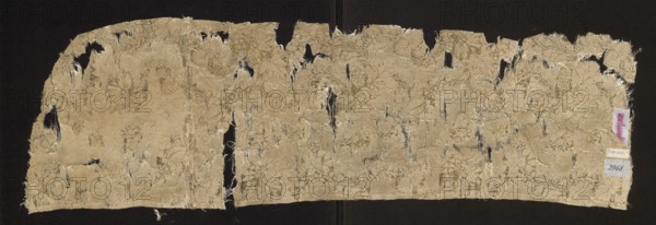 Fragment of silk fabric, patterned with plant tendrils and floral motifs in pink and yellow on a crimson background, 17th century, France; height 80 cm, width 23.5 cm., A wide, irregularly torn textile fragment with a floral woven pattern in beige and light gold tones against a dark background; the fabric shows frayed edges, several elongated holes, and areas of wear revealing darker backing through the openings, with a few small attached paper tags ignored and a narrow vertical seam visible where two pieces meet.