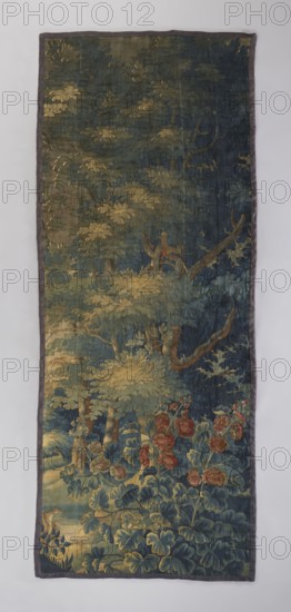 Fragment of verdure tapestry depicting flowers, leaves, floral ornaments, trees, birds, pastoral landscapes and interior furnishings; densely occupied surface with tree branches, and at the bottom tall plants with broad heart-shaped leaves; a crested bird hoopoe visible in the foliage; no border. Colors: navy blue, blue, green, and beige. 17th–18th century, Flanders. Wool tapestry, height 336 cm, width 136 cm., A vertical rectangular textile with a narrow dark border shows a lush forest scene dominated by deep greens and blues in the foliage and background, with areas of muted yellow and gold highlighting leaves and branches. In the lower section a group of large rounded leaves in bluish-green tones forms a dense undergrowth, punctuated by clusters of reddish flowers with warm orange and rust accents. Midway up, tree trunks and branches in brown and ochre rise through layered foliage, and a few paler yellow leaves catch light against the darker canopy. Upper areas are filled with overl...