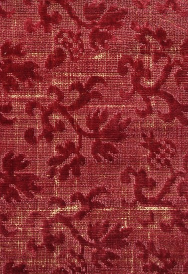 Fabric fragment, fragment of silk fabric chiseled velvet with floral pattern. Construction: basic warp and weft with beige silk thread, plain weave; pile with cherry-red silk weft — looped in the background, pattern raised above, sheared. Pattern repeat 0.7 cm x width 8.5 cm. Design: alternating small sprigs of several types arranged with vine and oak leaves and pomegranates or mushrooms, offset in rows by half the pattern width. Dated 1600–1630 after Markowsky; 1st half of the 17th century, Italy. Materials: silk; dimensions: height 16 cm, width 24 cm., A textured fabric with a raised floral and scroll pattern in deep red against a background of slightly lighter red with visible woven texture, with a few areas showing subtle golden highlights; the raised parts appear velvety while the background looks more coarse, and the overall colors visible are shades of red and touches of gold.