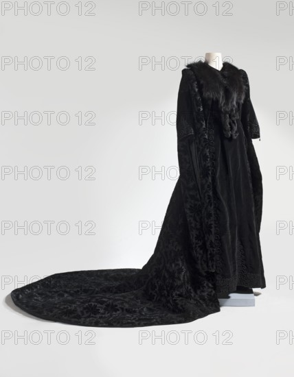 Dress - Helena Modrzejewska's stage coat. Coat of black patterned velvet, long, opens at the front, with a semicircular train. Fitted at the top, slightly flaring at the bottom. Very long sleeves reaching to the hem, slit at the top from the shoulder to mid-length, emphasized with a roller. Sleeves and edges lined with black silk, edges trimmed with drawstring. Black fox collar cut into small teeth on the neckline. 2nd half of the 19th century. Europe/United States place of origin uncertain. Women's garment, length 228 cm. Gift; provenance: Modrzejewska, Helena 1840–1909. Theater item; souvenir., A long black velvet garment displayed on a headless mannequin with a flowing train spread on the floor, featuring a dark fur collar and textured patterned velvet panels, with visible folds and drape in shades of black and deep charcoal against a pale gray background.