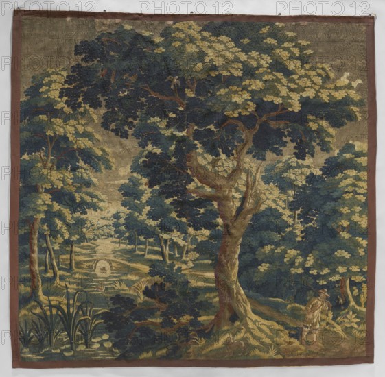 Tapestry — verdure., Subject: in the foreground a large tree with leafy branches filling the upper part of the fabric; on the right a hunter with a rifle; on the left a river with swimming ducks and a small bridge; floral ornaments and landscapes. Materials/technique: woven decorative fabric, tapestry. Colors: beige, green, blue. Date: 18th century. Place of origin: Flanders. Dimensions: height 250 cm, width 250 cm. Keywords: ducks, flowers, hunters, interior furnishings, weaving, verdure, decorative fabrics., A woven scene depicting a wooded landscape with a large central tree with a twisted trunk and spreading branches, surrounded by a group of smaller trees and dense foliage; the leaves appear in shades of dark blue, deep green, and pale yellow, while the tree trunks and ground show brown and tan tones; a winding path or clearing leads into the distance with grassy areas in muted green and yellow, and a small body of water reflecting lighter tones; in the lower right a solitary figu...