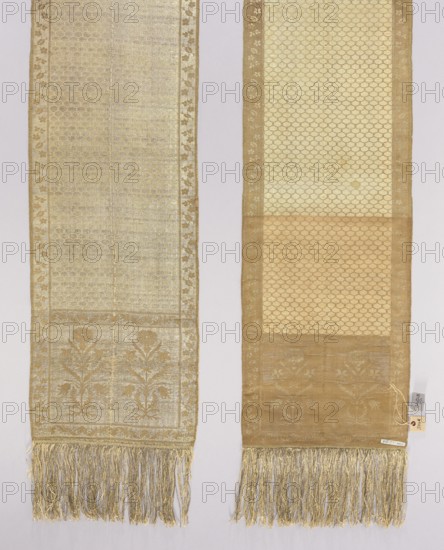 Kontusz sash, Paschalis Jakubowicz Manufactory Warsaw; belt manufacturer; 1788–1794. Bipartite heads with floral bouquets. Still with carp scale. Border with floral and plant twig. Sewn-on fringes. Ornament - Heads: two motifs of rooted flower bushes with a carnation flower at the top. - Center: with carp scale motifs. - Edges: vertical with motifs of flower branches forming a twig; horizontal with a twig motif with palmettes. - Borders: checkerboard. Coloring - Right: silver background, light brown ornament of heads and edges, cream carp scale design. - L.: reverse of right. Signed at the bottom in the heads: Paschalis. 18th century 1788–1790. Lipków, Warsaw . National costume, costume accessory. Length 372 cm, width 29 cm., A pair of long woven fabric panels hung side by side against a pale background, each panel narrow and rectangular with fringed ends; the left panel is a muted pale gold with metallic sheen and a subtle repeating scalloped pattern throughout, a floral border runnin...