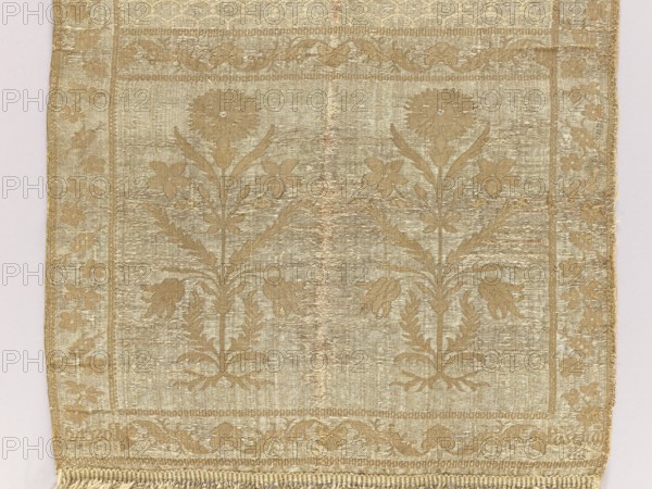 Kontusz sash by Paschalis Jakubowicz Manufactory Warsaw; belt manufacturer; 1788–1794. Bipartite heads with floral bouquets. Still with carp scale ornament. Border with floral and plant twig. Sewn-on fringes. Ornament - Heads: two motifs of rooted flower bushes with a carnation flower at the top. - Center: carp scale motifs. - Edges: vertical motifs of flower branches forming a twig; horizontal twig motif with palmettes. - Borders: checkerboard. Coloring - Right: silver background, light brown ornament of heads and edges, cream carp scale design. - Left: reverse of right. Signed at the bottom in the heads: Paschalis. Date: 18th century 1788–1790. Place of origin: Lipków, Warsaw . Type: national costume accessory kontusz sash. Dimensions: length 372 cm, width 29 cm., A rectangular woven textile with a repeating floral pattern in shades of gold and beige, showing a symmetrical design of stylized flowers with tall stems and leaves against a slightly lighter background, framed by a decorat...