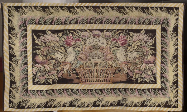 Kilim, 1st half of the 19th century. Wool fabric, height 123 cm, width 214 cm. Poland / Vilnius region, Lithuania. Biedermeier style. Interior decorative fabric court kilim. Entire main field filled with a motif of a basket with flowers and leaves, surrounded by a wide three-band border: widest band with a garland of leaves and flowers, two narrower bands with a diagonal pattern. Plant ornaments, flowers; colors: brown background with light and dark red, yellow, beige, and green ornamentation., A rectangular woven textile with a central dark field featuring a symmetrical floral arrangement in a basket, surrounded by multiple decorative borders; visible colors include dark brown, cream, pale yellow, green, pink, mauve, and beige, with touches of light blue and reddish tones. The central motif shows a basket filled with stylized flowers and foliage in cream, pink, mauve, green, and beige against the dark background. Around the central panel is a striped frame of pale yellow and cream, th...