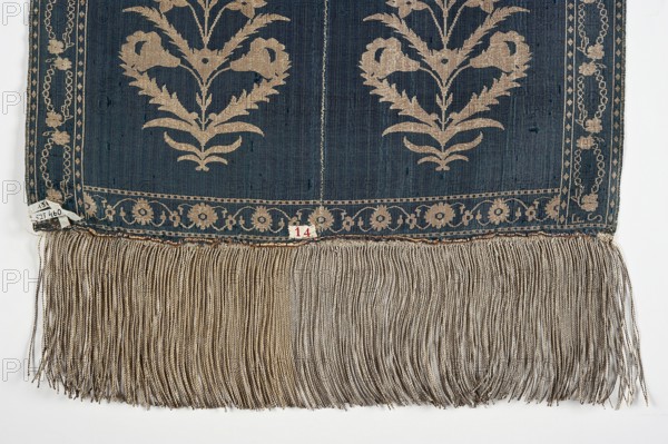 Kontusz belt, flower bushes ornament, double-sided belt, plant ornaments, Polish costume, Poland culture, half-solid belt, bipartite heads with an ornament of stylized flower bouquets, plain belts and belts with serrated ribbon, wreath edging. Colors: gold, silver, blue, black. 2nd half of the 18th century, Lyon, clothing accessory, national costume, length 360 cm, width 35 cm., A textile with a dark blue background and beige floral motifs and borders, showing a symmetrical plant-like pattern and a decorative band near the lower edge, with a long beige fringe hanging from the bottom; the colors visible include dark blue, beige, off-white, and hints of brown.