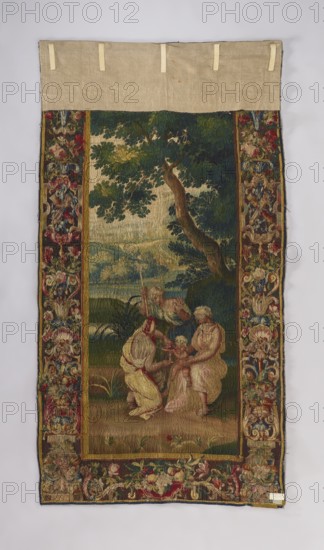 Figural tapestry, 18th century, Brussels. In the center of the landscape, little Dionysus kneels at the feet of one of the nymphs; another pours wine into his goblet, a third stands nearby with a spear. Surrounded by a border with motifs of flowers, acanthus leaves, shells, and ogee. Themes: flowers, floral ornaments, Dionysus, mythological scenes, mythological figures, figures of women with children, Bacchic scenes, nymphs. Materials/technique: weaving, decorative fabric for interior furnishings. Colors: brown, green, red, blue, ochre, sand. Dimensions: height 256 cm, width 136 cm., A vertical woven textile with a central scene bordered by an ornate floral frame, showing a group of figures beneath a large tree in a landscape; the top edge has a plain beige fabric extension with a few beige loops. The central area shows several human figures with pale skin tones wearing garments in shades of cream, pink, yellow, and muted red, positioned on green and brown ground with grass and small p...
