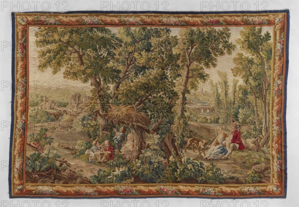 Figural tapestry, Chodecz Kuyavian-Pomeranian Voivodeship; flowers, floral ornaments, dogs, children, women, message provenance, genre scenes, genre scenes against a landscape background, tapestries, interior furnishings, pastoral scenes, decorative fabrics, tapestries. In the foreground a group of children playing against a background of tall trees. On the right a girl with a shepherd's staff sits on the grass and a young man next to them a dog. In the background on the right and left there is architecture. Narrow border with a motif of a simple frame entwined with flowers; cartouches in the corners. Colors: green, red, blue, yellow, brown, ochre, and cream. 2nd half 19th century, Germany; fabrics; height 265 cm, width 395 cm., A woven scene showing a pastoral landscape with a group of large trees with green and yellowish leaves dominating the center, a few figures seated beneath and beside the trees wearing clothing in muted red, blue, and cream tones, and a couple of animals nearby....