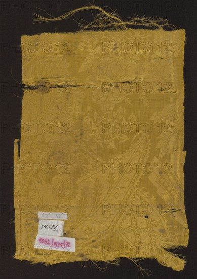 Fabric fragment, patterned silk damask, early 18th century, France. Yellow semi-silk damask with floral-fantasy motifs; background is warp-covered satin with a plain-weave ornament. Decorated with stylized large leaves with frayed edges, small heart-shaped leaves, small leaves and flowers, blades of grass, small fruit, details evoking Far Eastern architecture, and openwork lace-like elements in a bizarre style. Color: Jaune d'or Newton. Pattern scale larger than preserved fragment. Dimensions: height 34 cm, width 23.5 cm., A rectangular piece of worn fabric with frayed edges and loose threads displayed against a dark background, the fabric showing an intricate woven floral and geometric pattern visible through texture and subtle sheen, with areas of thinning and small holes and a horizontal tear near the top; the dominant color of the fabric is golden yellow with variations in tone, and there are a few small labels attached near the lower left corner that contain handwriting and printe...