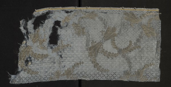Fabric fragment, fragments of silk patterned fabric, report composition based on two intersecting diagonal arrangements. One arrangement formed by ribbons winding wavy-like from tiny fancy leaves brocaded with silver thread, the other running opposite with forms resembling sheaves brocaded with gold thread. Satin background enriched with a miniature rhomboidal net pattern in liseré. Blue Gros de Tours background with elements of liséré. Brooch motifs: silver lamellar and gold-braided thread with a loose braid showing the silk core in natural color. Report size larger than preserved fragment. Motifs in the bizarre fabric type. Multicolored border: green, pink, and yellow threads. Late 17th century, France. Height 56 cm, width 27 cm., A torn rectangular piece of fabric with frayed edges rests against a dark background, showing a woven floral and foliage pattern; the fabric is primarily pale bluish-gray with accents of muted gold and silvery threads, and there are several large holes and ...