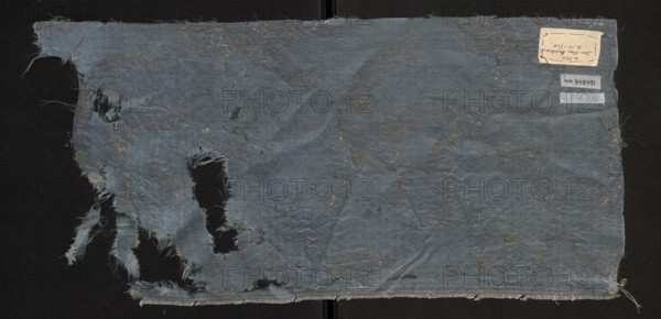 Fabric fragment, fragments of silk fabrics, patterned fabrics. Report composition based on two intersecting diagonal arrangements: one formed by ribbons winding wavy-like from tiny, fancy leaves brocaded with silver thread; the other running in the opposite direction with forms resembling sheaves brocaded with gold thread. Satin background enriched with a miniature rhomboidal net pattern in liseré. Blue Gros de Tours background with elements of liséré. Brooch motifs: silver lamellar and gold-braided thread with a loose braid showing the silk core in natural color. Report size larger than the preserved fragment. Motifs in the bizarre fabric type. Multicolored border: green, pink, and yellow threads. Late 17th century, France. Height 56 cm, width 27 cm., A worn rectangular piece of fabric laid flat with frayed edges and several irregular holes, showing loose threads and torn strips along one side; the fabric is primarily a muted blue with patches of darker blue where folds and wear appea...