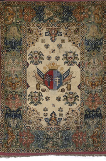 Rug with the Potocki and Mniszech coats of arms. A light central field surrounded by a wide three-band border; the widest band has a palmette motif. In the central field is an octagonal coat-of-arms cartouche: on the right the Pilawa coat of arms on a blue background, on the left the Konczyce coat of arms on a red background; above the cartouche a noble crown; behind the cartouche two crossed hetman's maces and also swords, pikes, banners, and cannon barrels. Below the cartouche are symmetrically arranged pairs of barrels and cauldrons, and two stacks of cannonballs. Possibly 1st half of the 18th century, Brody, Poland. Fabrics; height 209 cm, width 142 cm., A rectangular woven textile with a central cream field surrounded by a wide patterned border, featuring a central shield motif in red and blue topped by a gold crown and flanked by stylized banners in blue and tan; the cream field contains several floral rosette motifs in blue, tan, red, and green and pairs of symmetrical stylized ...